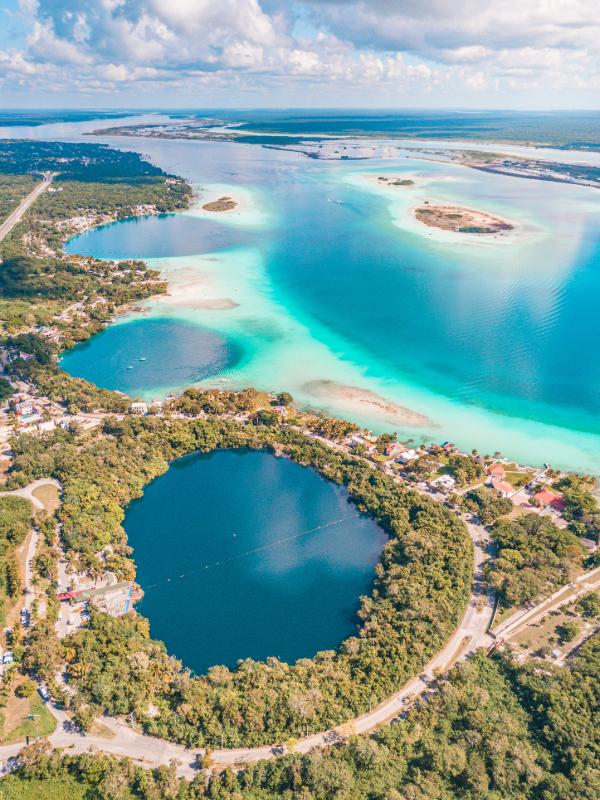 A stunning view of Cenote Azul, a large open-air cenote with crystal-clear turquoise waters, surrounded by rocky edges and lush greenery, located between Playa del Carmen and Tulum, Mexico. One of the most magical Tulum Cenotes.