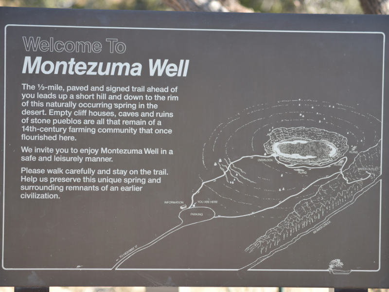 "A welcome sign at Montezuma Well, cautioning visitors to stay safe, set against the backdrop of the limestone sinkhole, lush vegetation, and ancient Sinagua cliff dwellings."