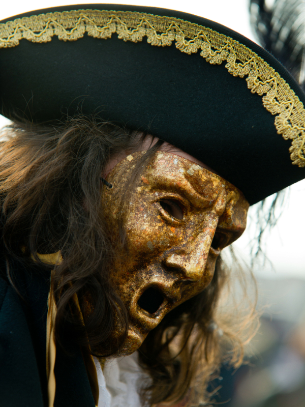 An actor wearing a goldish, weathered Trojan War chorus mask, dressed in 1700s English attire. The mask features exaggerated facial features, blending ancient Greek theater with 18th-century fashion.