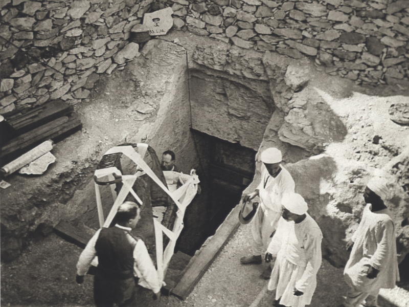 A historic 1922 photograph by Harry Burton showing archaeologists carefully transporting the chariot body discovered in King Tutankhamun's tomb. The chariot, an important funerary artifact, reflects the luxury and complexity of the burial items placed in the tomb.