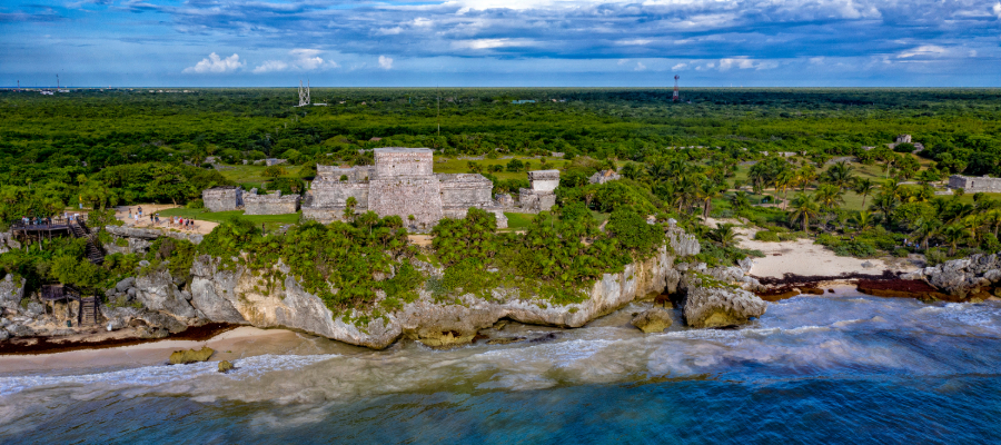 A panoramic aerial view of the Tulum ruins perched on a cliff overlooking the blue Caribbean Sea, surrounded by lush rainforest canopy, l and stretching coastline. Tulum Cenotes vary in location.