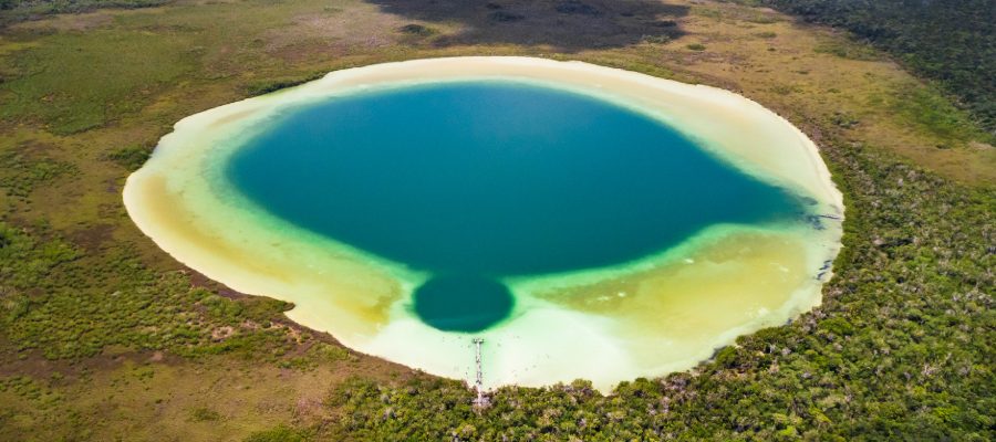 A stunning aerial view of Laguna de Kaan Luum near Tulum, Mexico, showcasing its turquoise shallow waters, a deep cenote at the center, and surrounding lush rainforest.