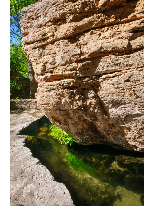 "Montezuma Well, a natural limestone sinkhole near Camp Verde, Arizona, within Montezuma Castle National Monument, displaying a stream carving through the rock wall alongside a walking path."