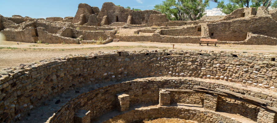 "Panoramic view of the Aztec Ruins National Monument in New Mexico, highlighting the ancient Puebloan stone structures, interconnected rooms, and a visitor walkway, set against an arid desert landscape with distant mountains."