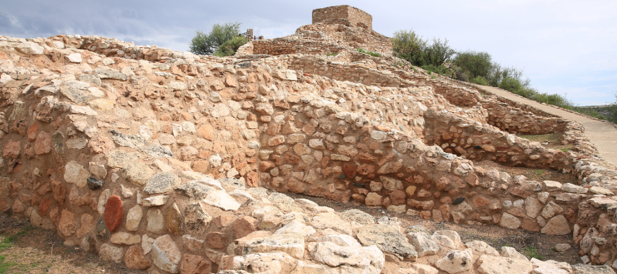 "The image shows Tuzigoot National Monument, featuring the ancient Tuzigoot Pueblo ruins in Arizona. The multi-level stone structure, built from local materials, sits atop a hill with a smooth, well-maintained pathway winding around and up to the ruins. The clear blue sky and sparse desert vegetation, including shrubs and trees, highlight the arid Arizona landscape. The stonework displays varied colors and textures, reflecting the natural materials used in construction. The scene evokes a sense of history and the ancient culture of the Sinagua people."