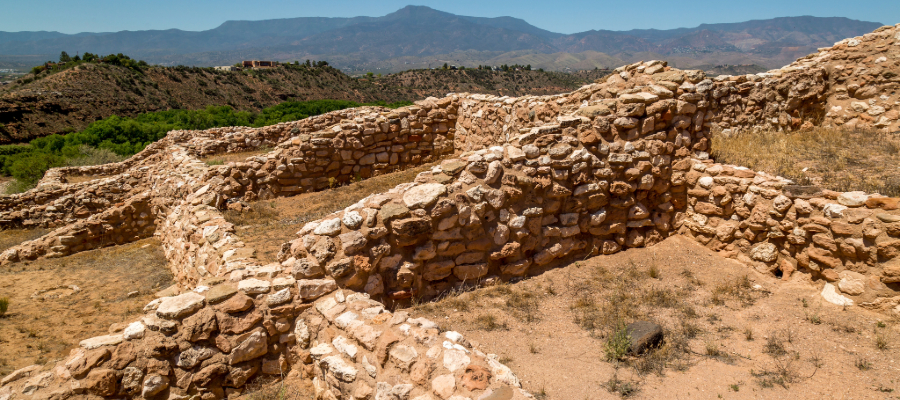 Tuzigoot National Monument, an ancient Sinagua pueblo ruin, perched on a hilltop with a stunning view of the Verde Valley and distant mountains in the background. The stone structures stand against a dramatic landscape of desert vegetation and rugged peaks
