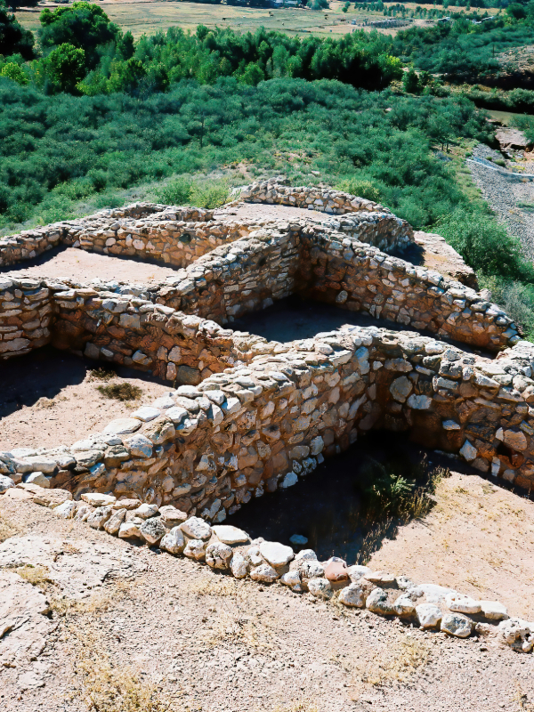"The image shows the ruins of Tuzigoot National Monument, also known as Tuzigoot Pueblo, located near Clarkdale, Arizona. The ancient Sinagua village, built on a hilltop, overlooks the Verde River valley. The stone and mortar structure features multiple rooms and levels, showcasing the architectural style of the Sinagua people. The surrounding landscape includes desert vegetation and distant hills, highlighting the arid beauty of the Arizona environment."
