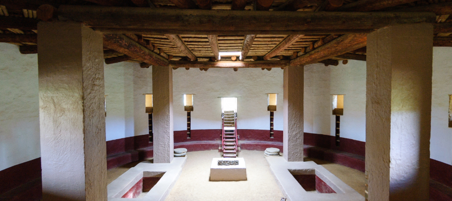 "The Great Kiva at Aztec Ruins National Monument, a large circular ceremonial structure made of sandstone blocks, partially underground with a roof supported by wooden beams, set against an arid landscape and clear blue sky."