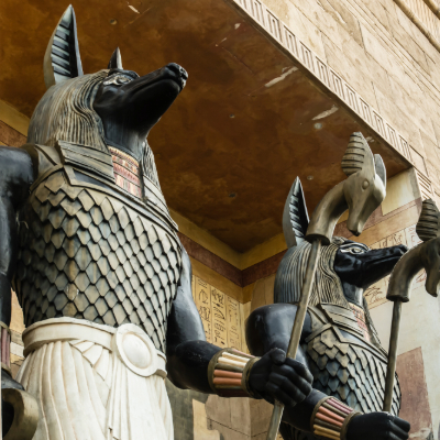 "Twin statues of Anubis, the jackal-headed Egyptian god of death and mummification - shown in detailed ceremonial attire with scale-patterned collars and staffs, possibly in a museum or theme park setting"