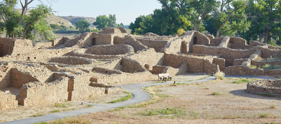 "Aztec Ruins National Monument in New Mexico, featuring well-preserved ancestral Puebloan stone and adobe brick structures, interconnected rooms, and a paved walkway for visitors, set in an arid landscape with sparse vegetation and distant hills."