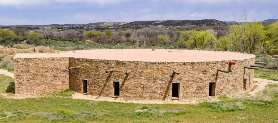 "The Great Kiva at Aztec Ruins National Monument in New Mexico, a large circular ceremonial structure made of sandstone blocks, partially underground with a roof supported by wooden beams, set in an arid landscape with low hills and a clear blue sky."
