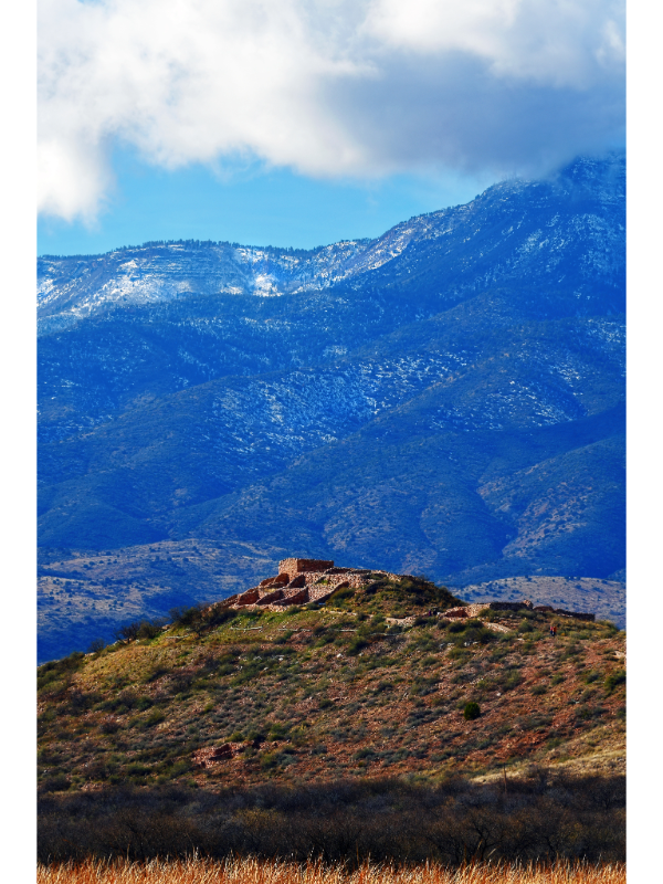 "The image shows Tuzigoot National Monument in Arizona, with the ancient pueblo ruins perched atop a hill. The foreground features a grassy slope leading up to the reddish-brown stone structures, which blend seamlessly into the landscape. In the background, a snow-dusted mountain range rises under a clear blue sky with scattered white clouds. The daytime lighting highlights the terrain's texture and emphasizes the monument's harmony with its natural surroundings."