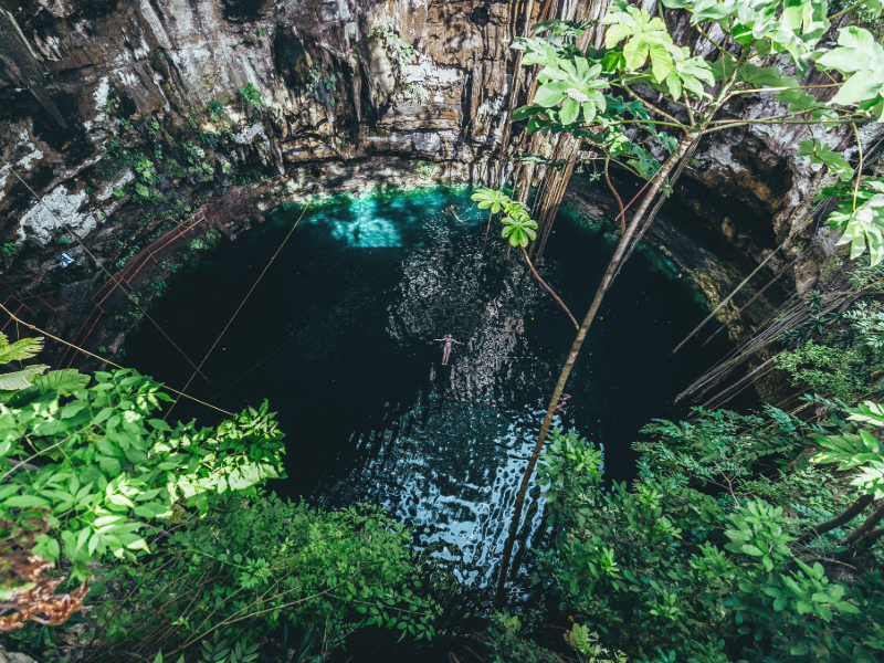 Tulum Cenote: A serene cenote surrounded by rocky walls and lush greenery at Hacienda Cenote Oxmán near Valladolid, Mexico, with a wooden swing rope and crystal-clear water.