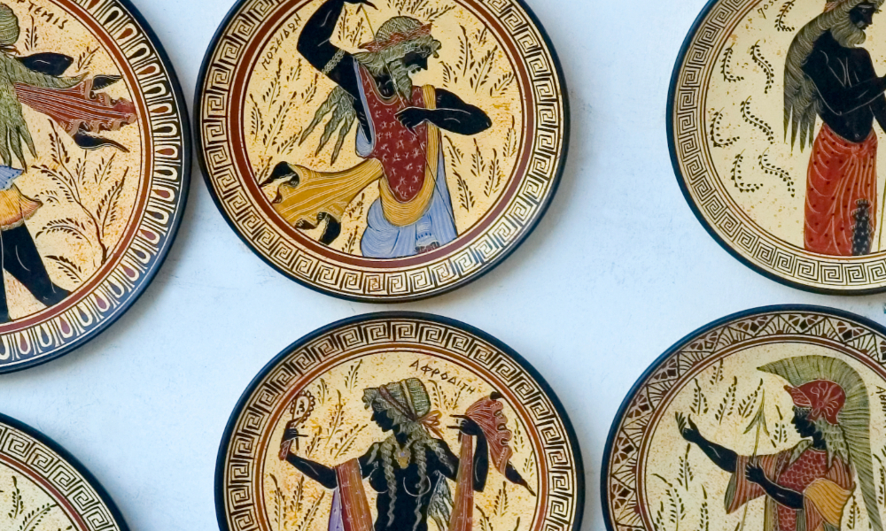 Ancient Greek black-figure pottery aka terracotta kylix (drinking cup) depicting Poseidon, Aphrodite and Themis in black silhouette against red clay background, with incised details and red/white accents.