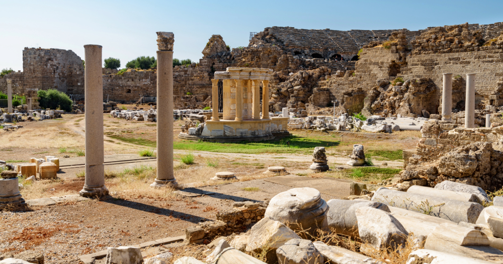 Ruins of the ancient agora in Side with scattered marble columns, temple remnants, and a well-preserved Roman theater in the background